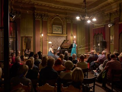Historischer Saal, zwei Damen im Abendkleid auf der Bühne, eine am Klavier, eine singt stehend, sitzendes Publikum