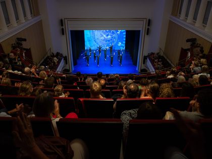 Blick aus dem Oberrang des Theaters, viele gefüllte Zuschauerreihen, die Bühne ist ganz in blau, darauf tanzen 11 einheitlich schwarz gekleidete Personen